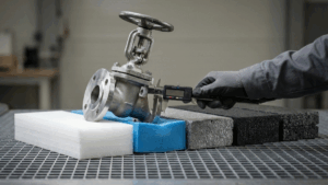 Análisis técnico de deformación: un ingeniero utiliza un calibre digital para medir la resistencia de muestras de espuma (PE y EPP) bajo el peso de una válvula industrial. La imagen muestra la comparativa de densidad y soporte de diferentes polímeros para la optimización de embalajes de protección en logística avanzada.