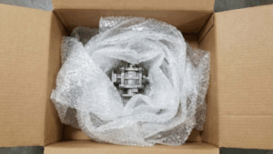 Pequeña pieza industrial de metal envuelta en una cantidad excesiva de papel de burbujas dentro de una caja de cartón grande, ejemplo de embalaje ineficiente.