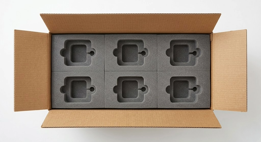 Una toma de estudio desde arriba mirando hacia una caja de cartón marrón abierta, que contiene seis insertos de espuma de polietileno gris perfectamente ajustados a los lados. Sin embargo, la caja es notablemente más alta de lo necesario ; hay un gran espacio vacío y desaprovechado entre la parte superior de los insertos de espuma y las solapas superiores de la caja. Esto ilustra el problema de diseño de embalaje con márgenes generosos o adaptación a estándares existentes , lo que resulta en un sobrecoste logístico al transportar y almacenar "aire" innecesario