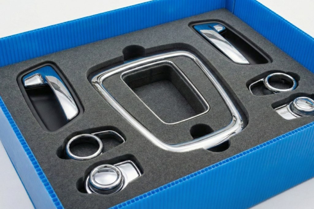 Vista en ángulo de una caja de plástico corrugado azul que contiene un inserto de espuma técnica gris oscuro con troquelado a medida. En los compartimentos se alojan diversas piezas de automóvil con acabado cromado brillante, incluyendo manetas de puertas, marcos de consola y mandos circulares. El diseño del embalaje resalta la organización y la protección individual de cada pieza para evitar arañazos durante el transporte.